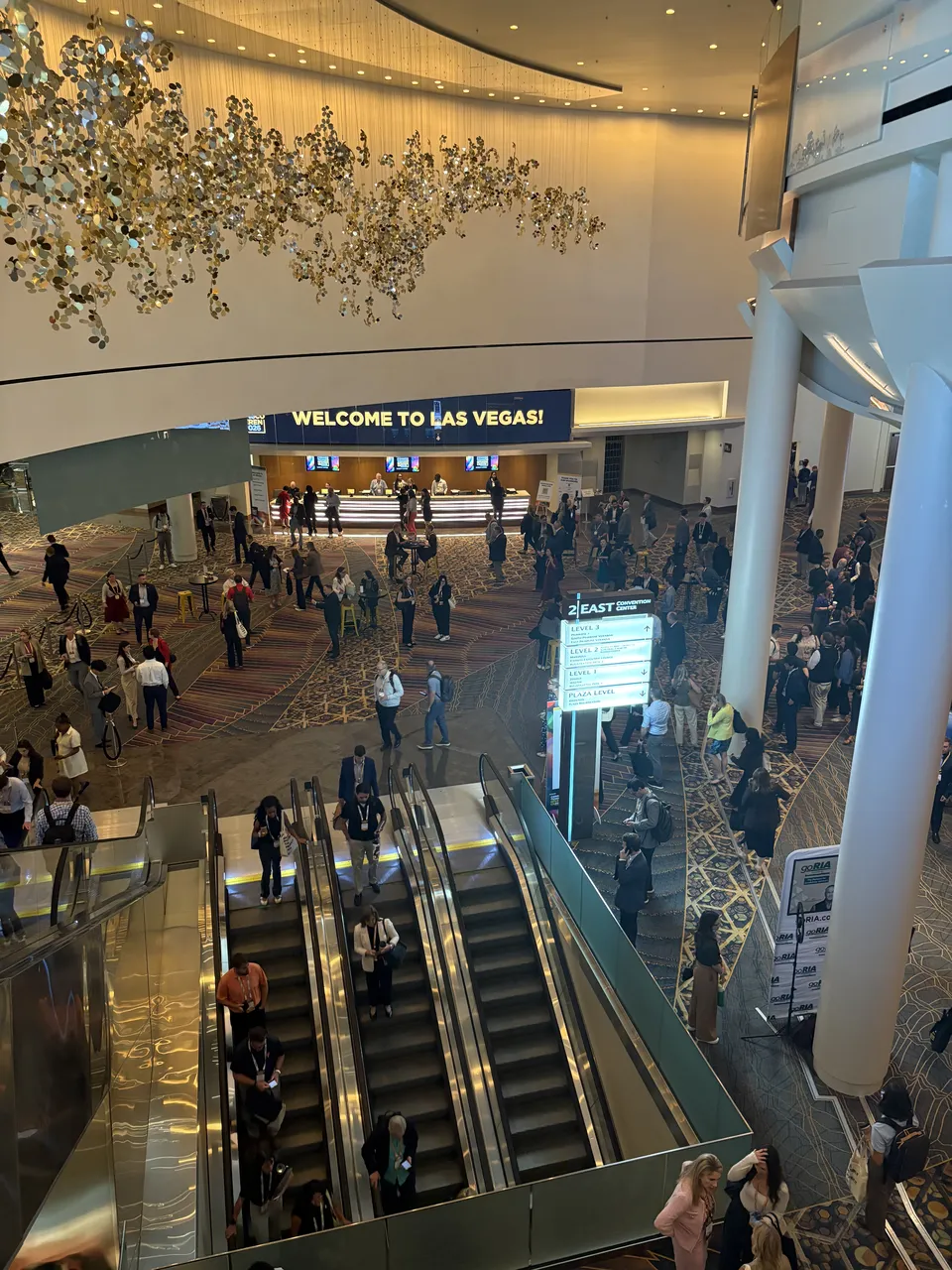 Aria 会议中心大堂，扶梯上下都是挂着胸牌的参会者，远处写着 Welcome to Las Vegas